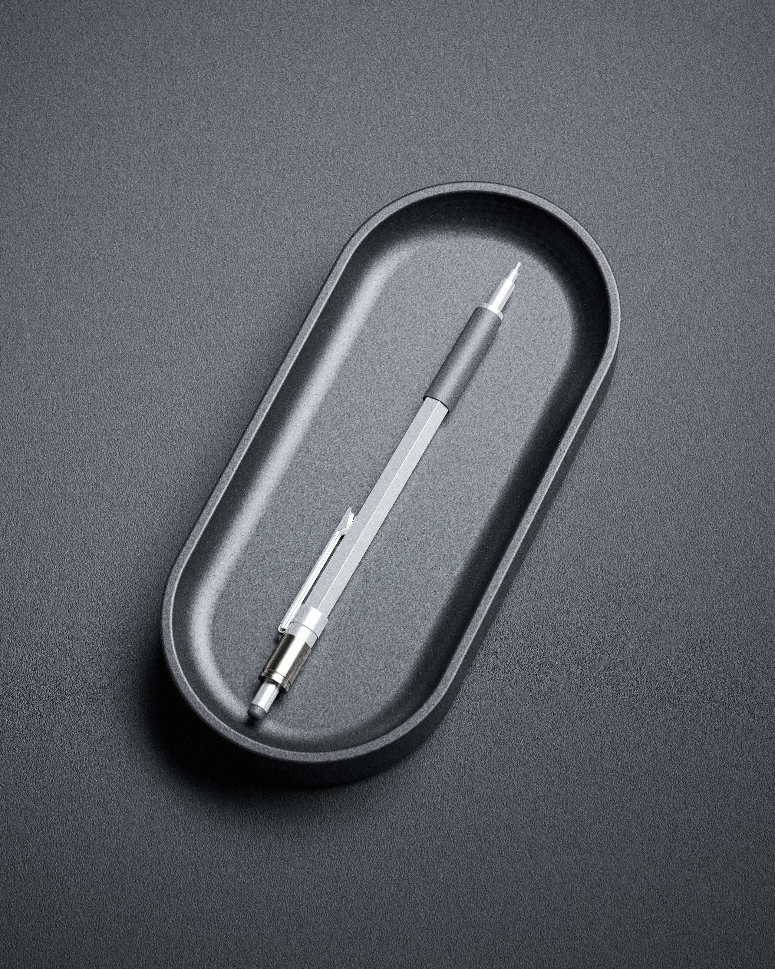 The Kuro Axis Capsule, a silver mechanical pencil with a black grip, rests in a sculptural pen tray on a textured gray surface, bringing subtle elegance to your workspace.
