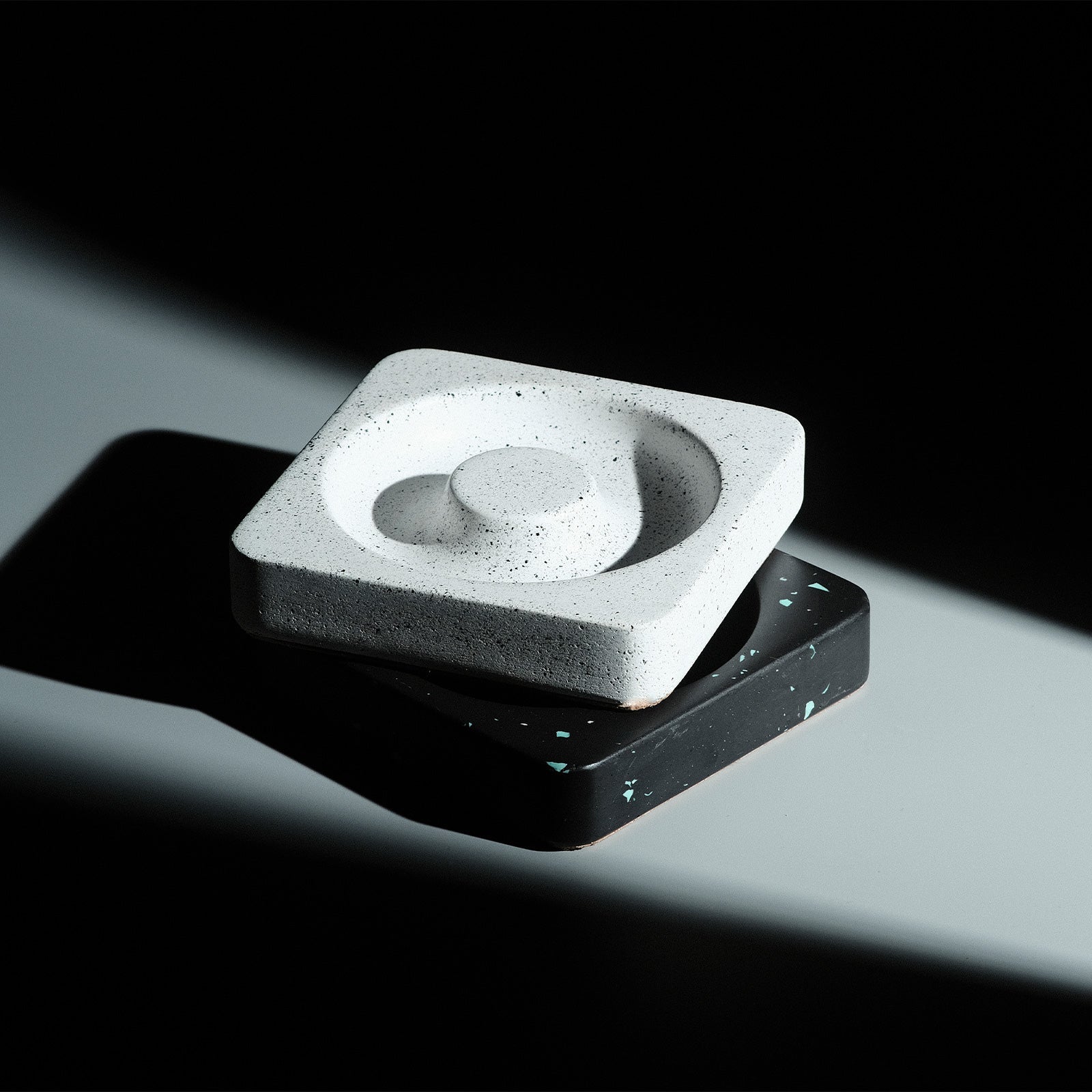 Minimalist black geometric trays and containers of various shapes and sizes are arranged on a smooth, light-colored surface with soft overhead light casting gentle shadows.