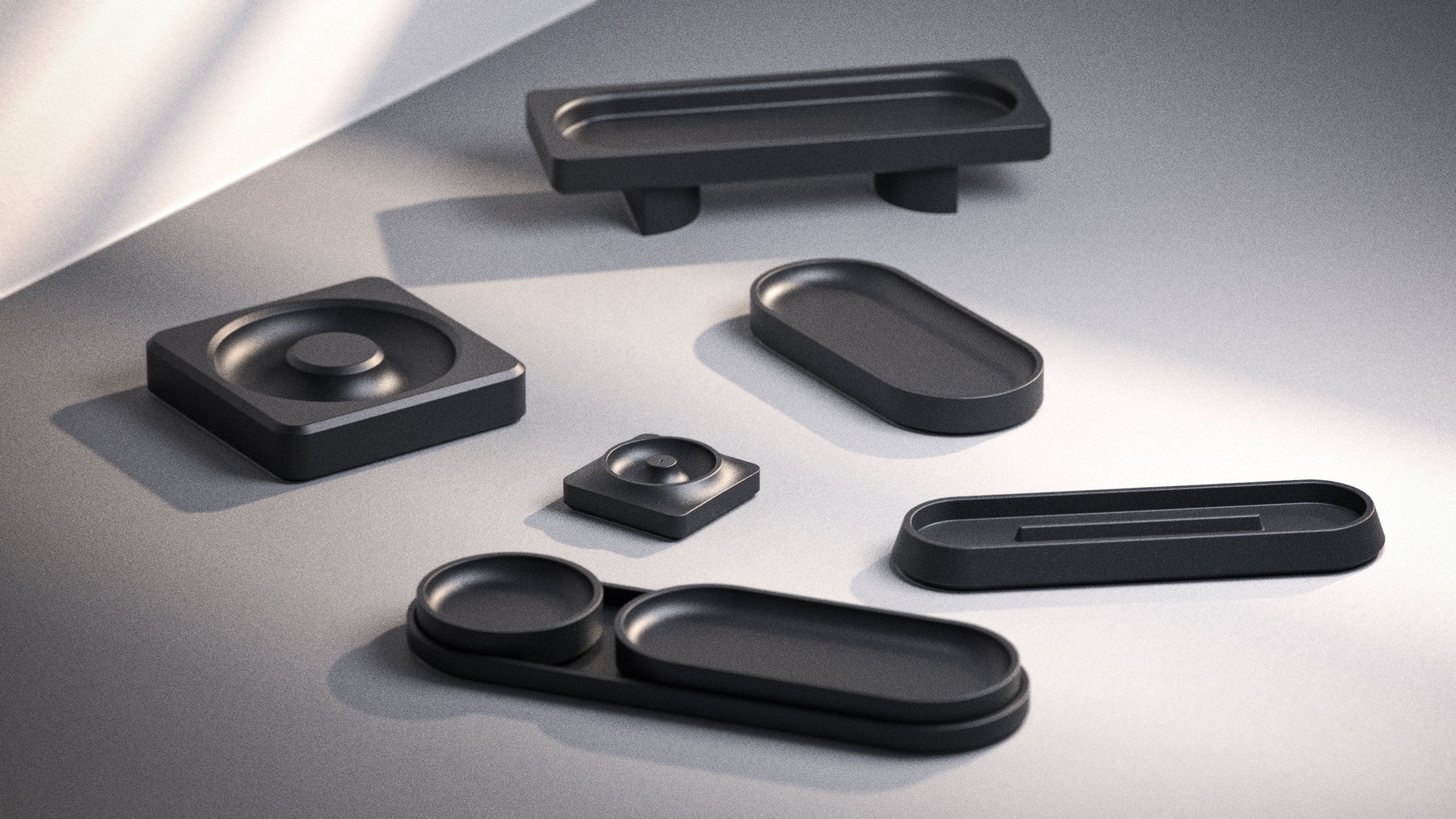 A collection of sleek, matte black geometric trays and containers of various shapes and sizes arranged on a smooth, gray surface with soft natural light.