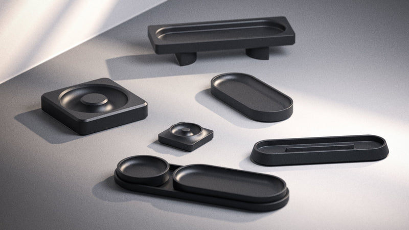 A collection of sleek, matte black geometric trays and containers of various shapes and sizes arranged on a smooth, gray surface with soft natural light.
