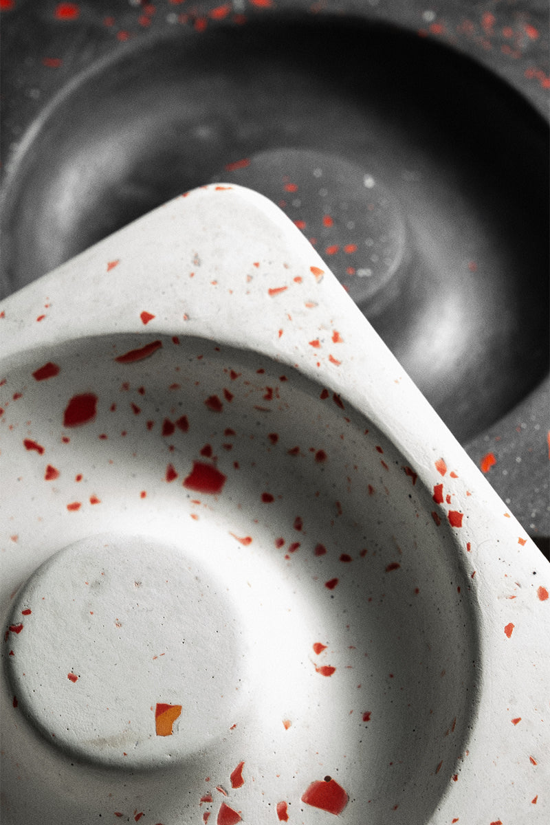Close-up of Kuro’s Ash Orbit: two partially overlapping circular trays—one white with red speckles, one dark gray—both with textured surfaces and a central indentation, creating a modern, donut-inspired sculptural ash tray centerpiece.