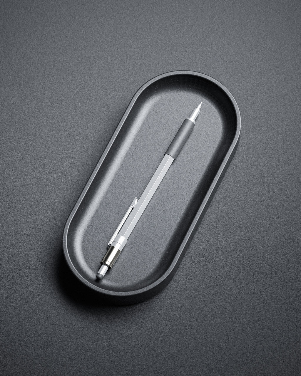 The Kuro Axis Capsule, a silver mechanical pencil with a black grip, rests in a sculptural pen tray on a textured gray surface, bringing subtle elegance to your workspace.